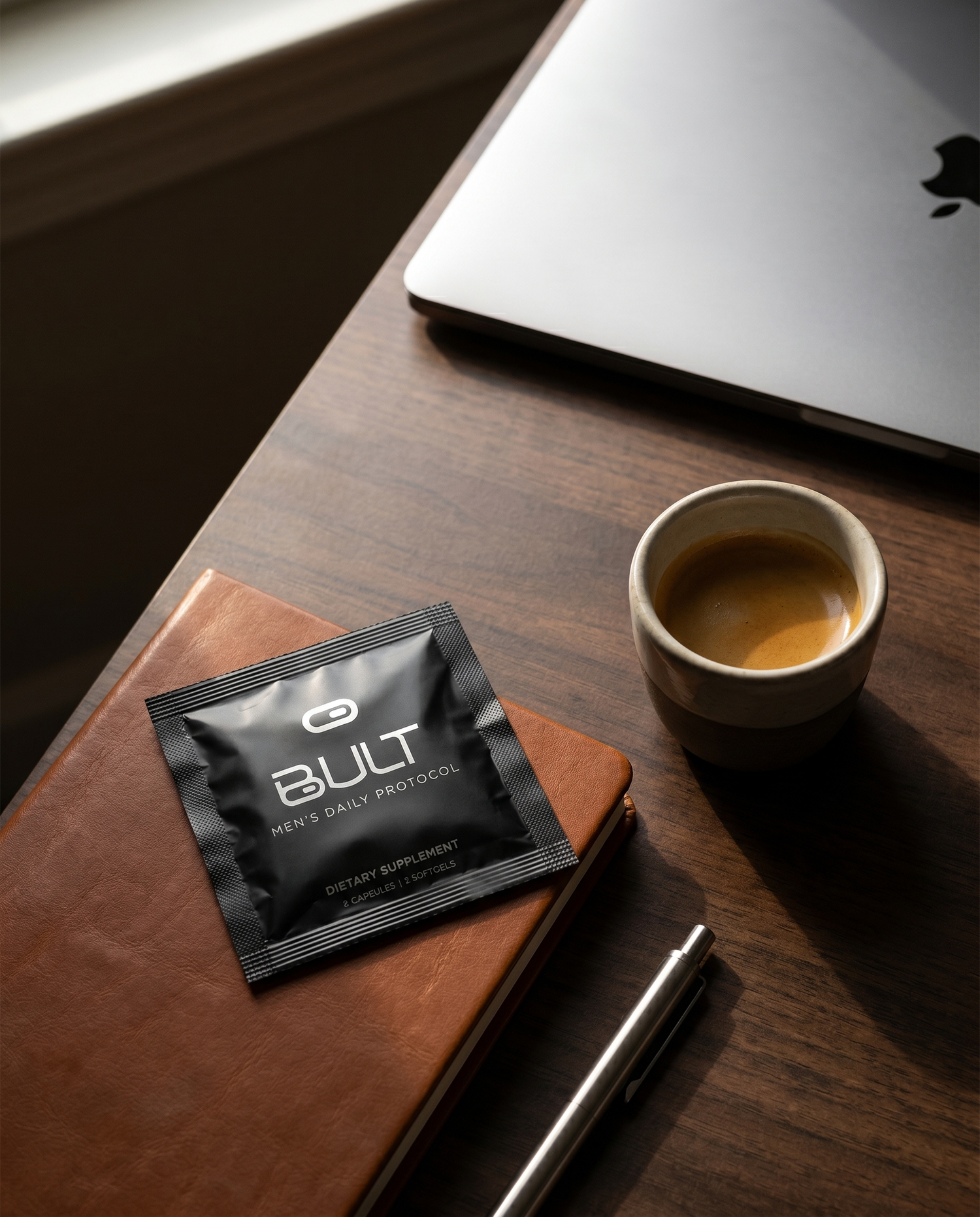Bult Men's Daily Vitamin Protocol supplement packet on a wooden desk with a cup of coffee and laptop.